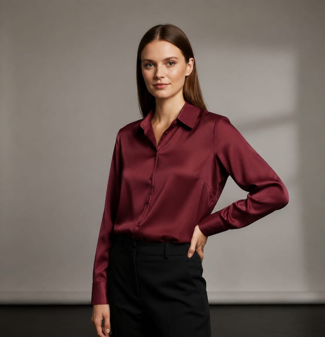 A woman modeling a long-sleeve satin button-up shirt in deep burgundy, paired with black trousers, against a neutral gray studio background.