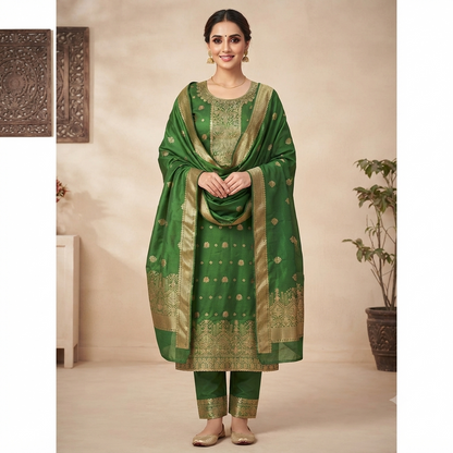 A woman models a green traditional ethnic outfit with gold embroidery, consisting of a kurta, pants, and a matching dupatta, set against a warm beige background with decorative wall art and a potted plant.