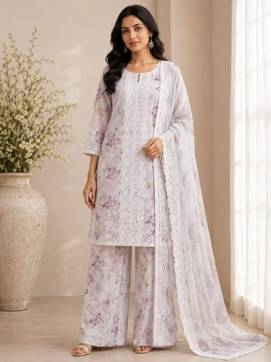 A woman models a traditional three-piece salwar kameez suit in a soft off-white color with delicate purple floral prints and intricate lace embroidery. The set includes a long-sleeve tunic, wide-leg trousers, and a matching sheer dupatta draped over her shoulder, set against a warm beige background with a large decorative vase of flowers.
