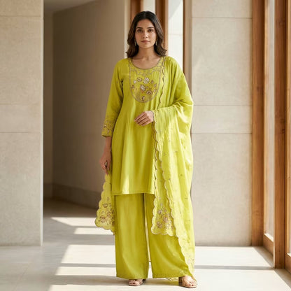 A woman models a bright yellow-green Indian ethnic suit set consisting of a kurta with floral embroidery, matching wide-leg trousers, and a dupatta with scalloped edges, standing in a modern architectural hallway with warm natural light. Jukarsh Fashion