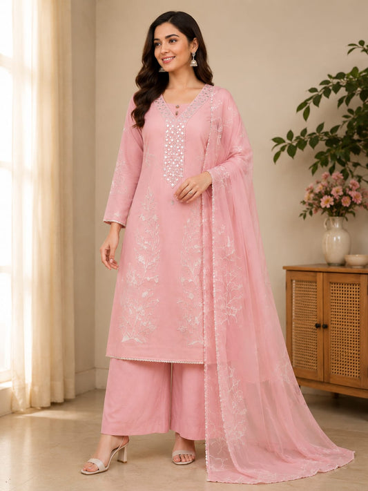 A woman models a light pink traditional Indian outfit consisting of a long kurta with white floral embroidery, matching wide-leg trousers, and a sheer dupatta, set against an indoor background with a wooden cabinet and potted plants.