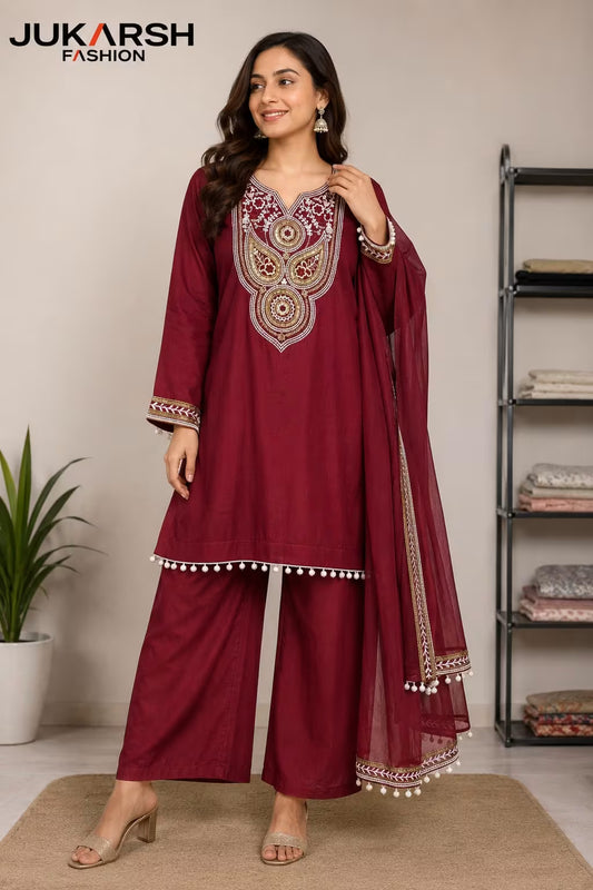 A woman models a three-piece salwar kameez suit in deep maroon. The set includes a long-sleeved kameez top with ornate gold and silver embroidery around the neckline, matching wide-leg trousers, and a sheer dupatta with embroidered borders and white pom-pom trim.