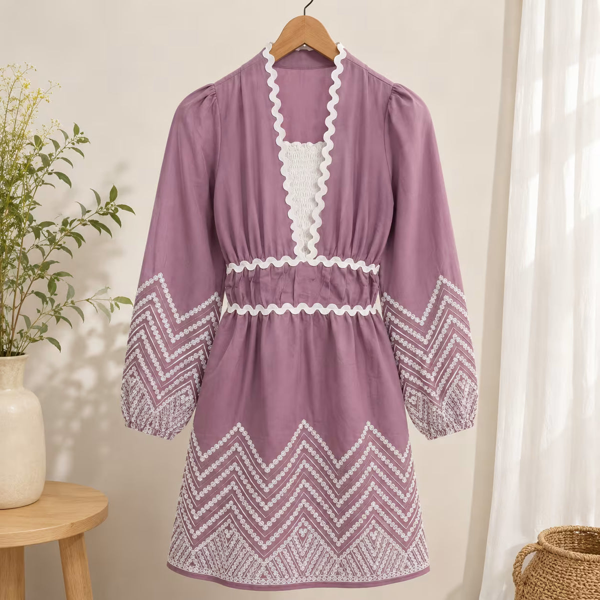 A mauve purple long-sleeve dress with white embroidered chevron patterns and a white lace V-neckline, hanging on a wooden hanger against a warm beige wall with soft window light.